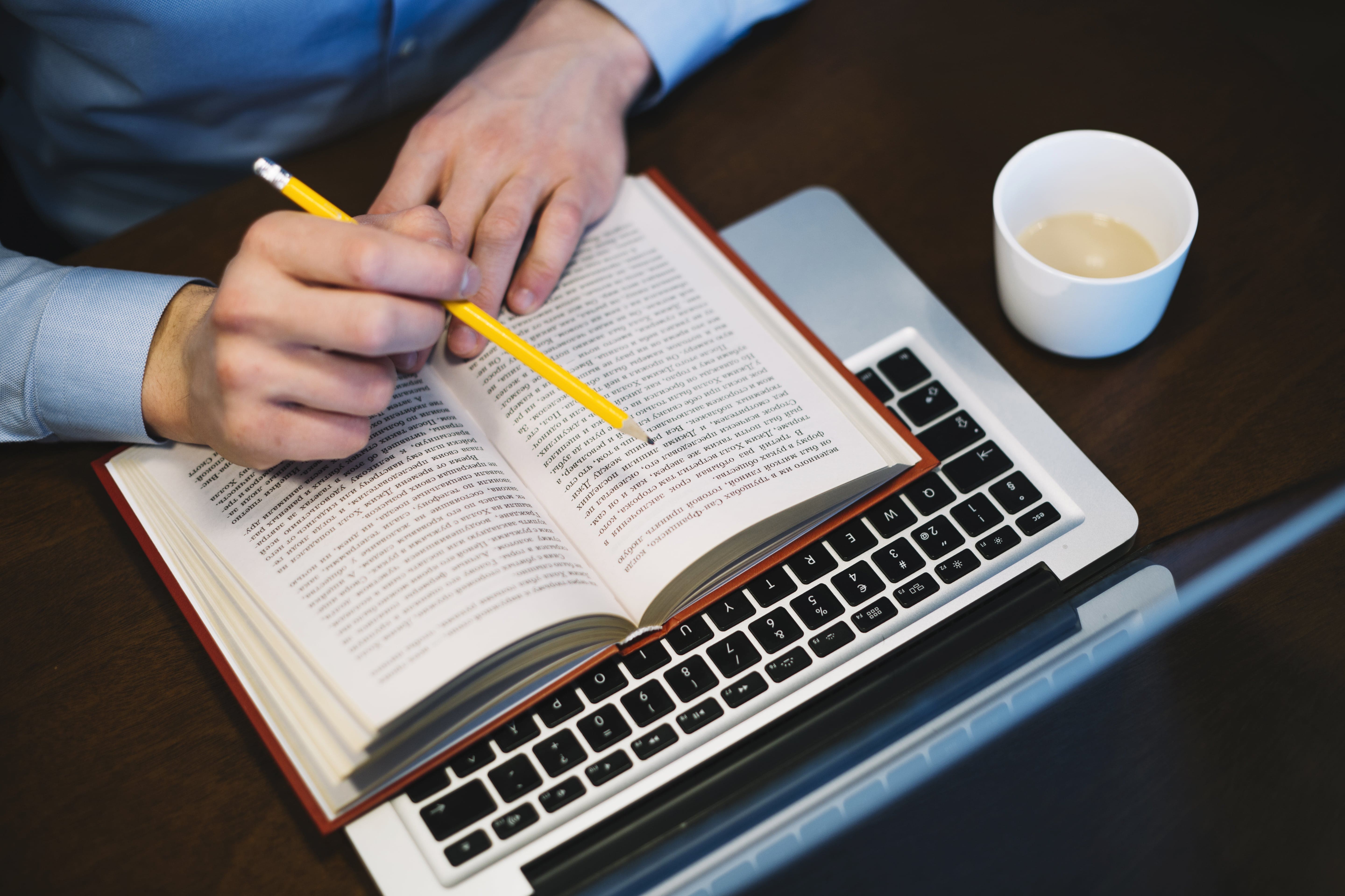A man writing on a book with a pencil and sipping coffee, engrossed in his work.